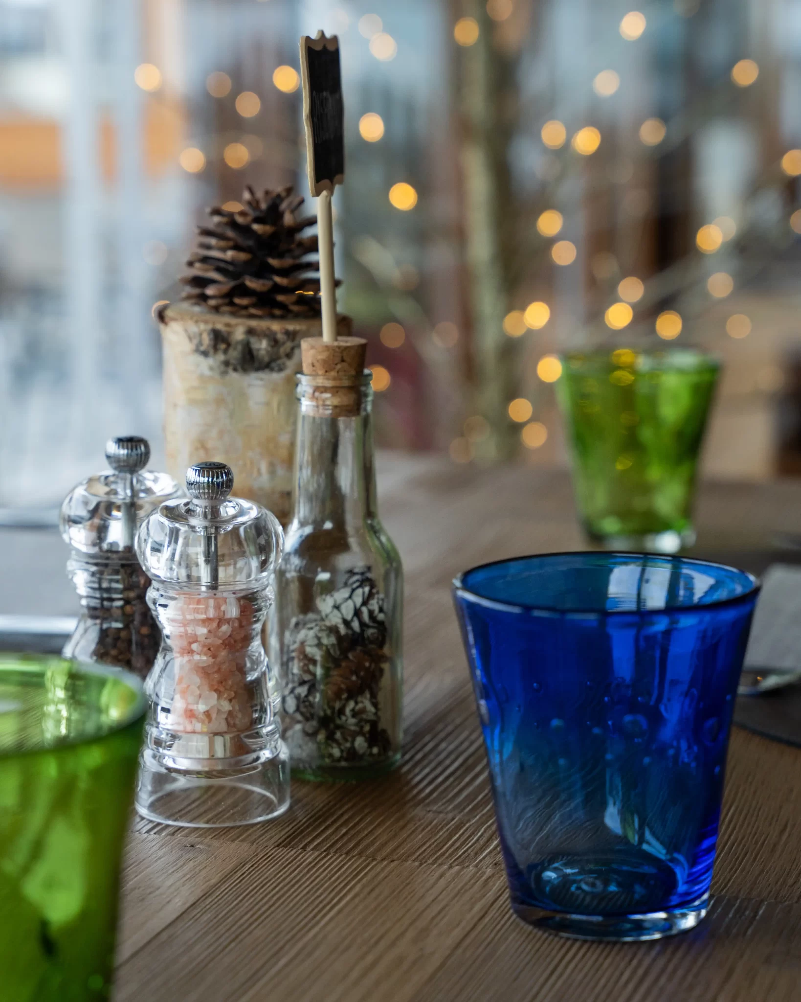 Bicchieri colorati e condimenti su un tavolo di legno con luci soffuse sullo sfondo.