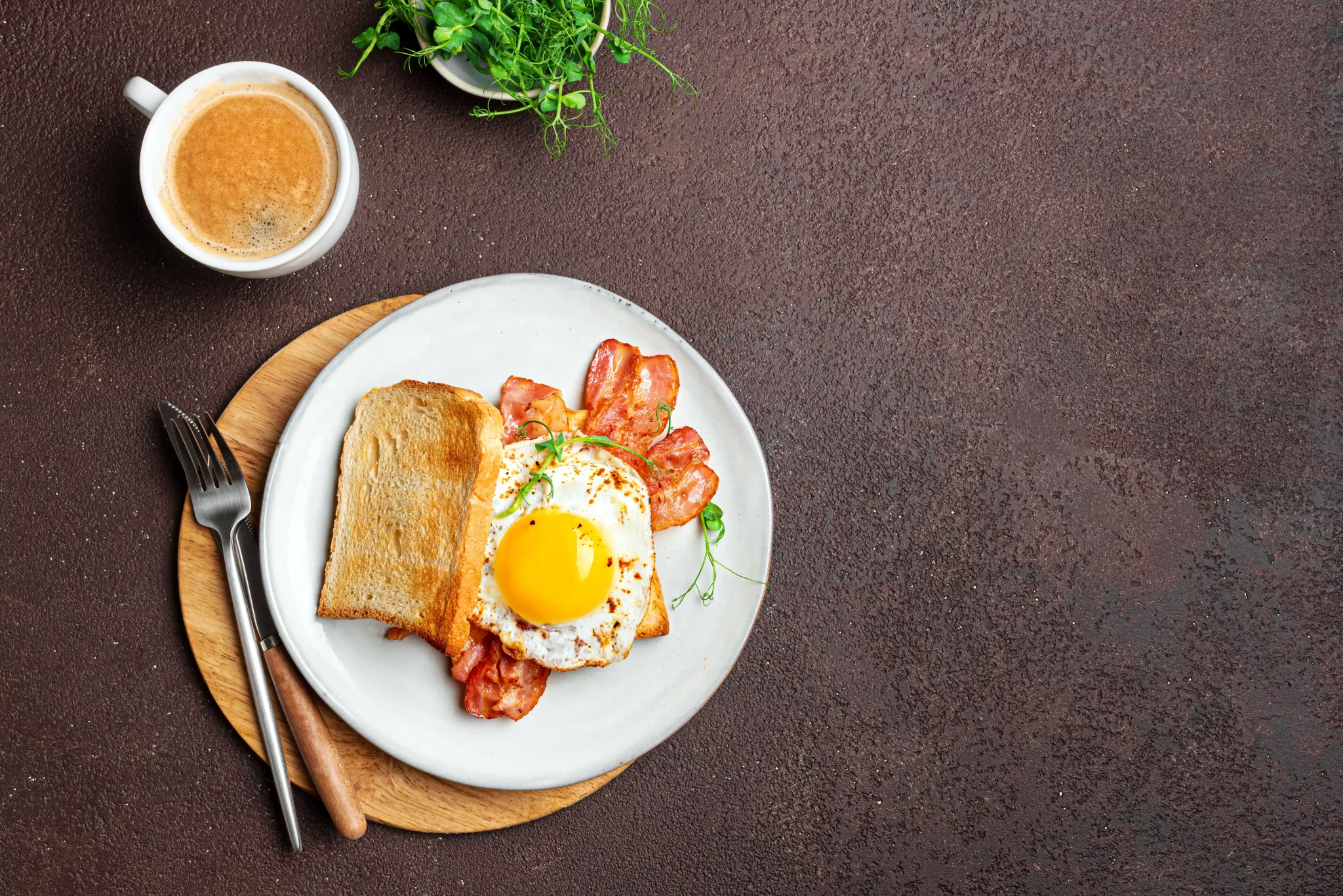 Colazione con uovo fritto, pancetta e pane tostato su un piatto accanto a una tazza di caffè.