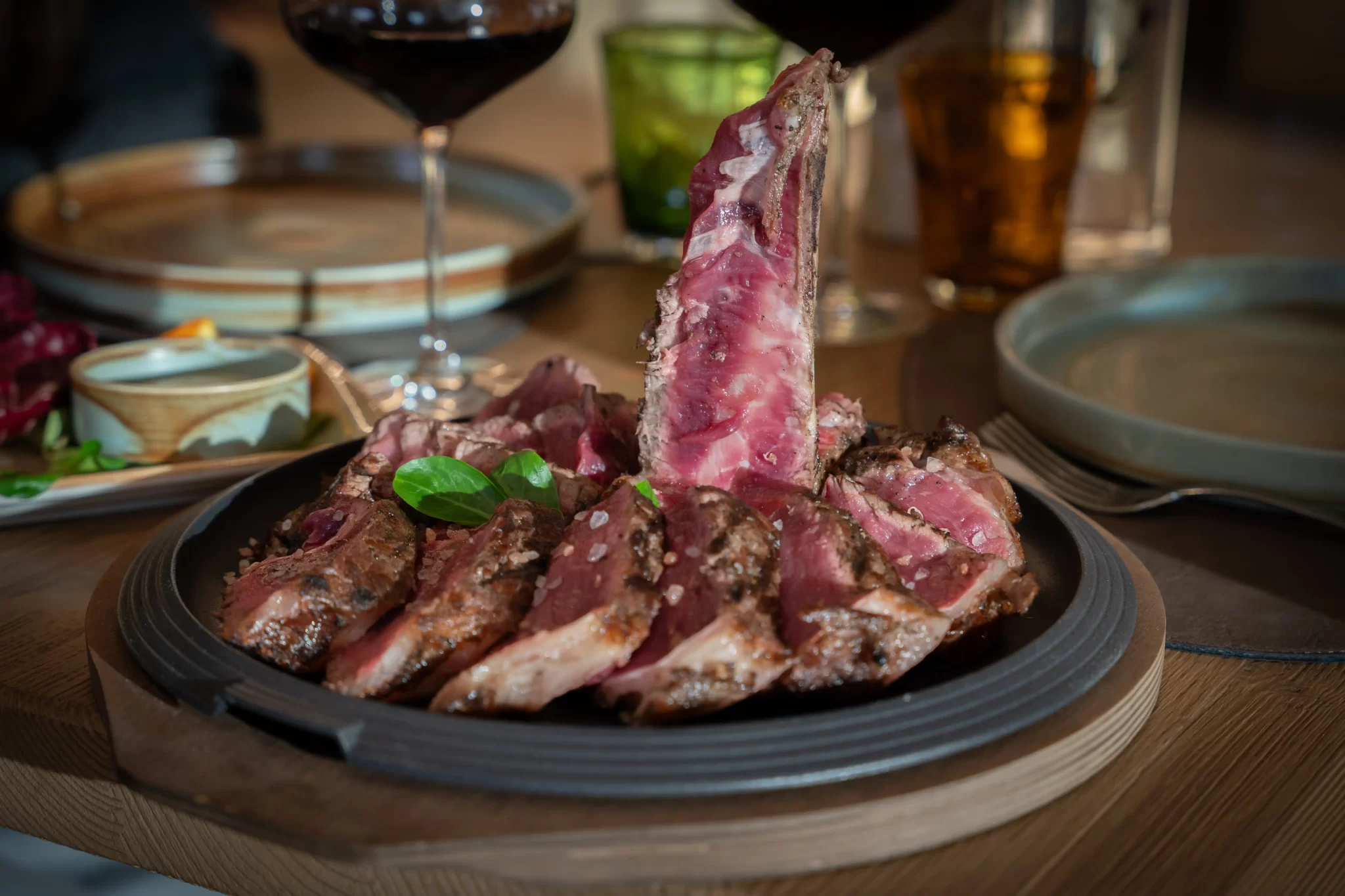Bistecca di carne grigliata su un piatto con contorni e bicchieri di vino sullo sfondo.