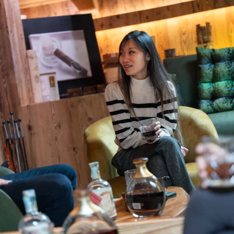 Una donna seduta in un salotto accogliente mentre tiene in mano un bicchiere di bevanda, circondata da bottiglie su un tavolino.