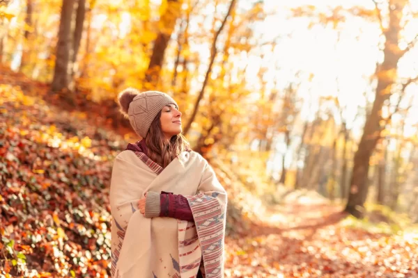 Donna sorridente avvolta in una coperta in un bosco autunnale soleggiato.