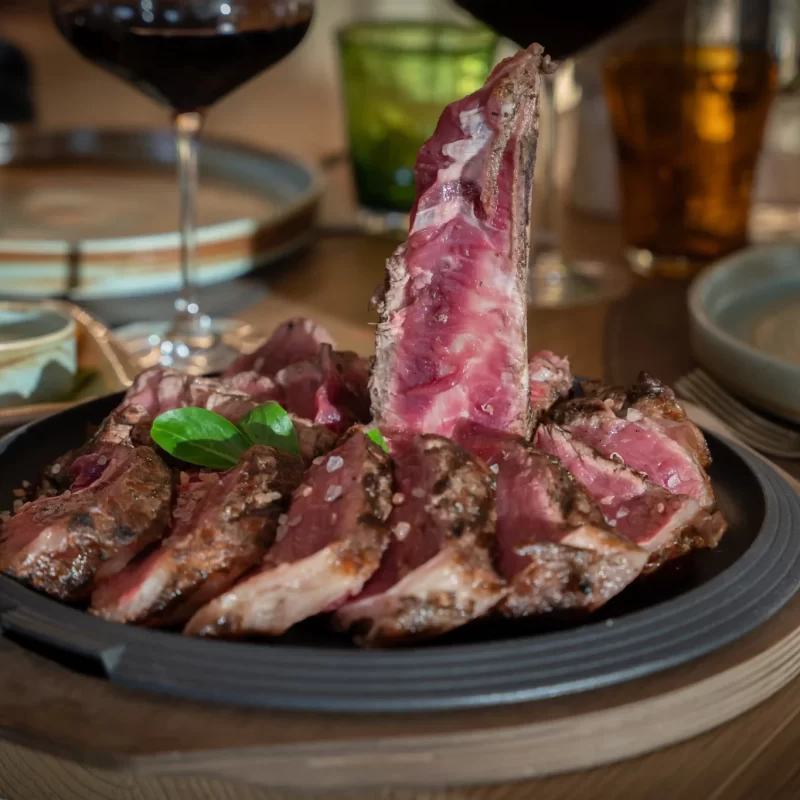 Bistecca di carne grigliata su un piatto con contorni e bicchieri di vino sullo sfondo.