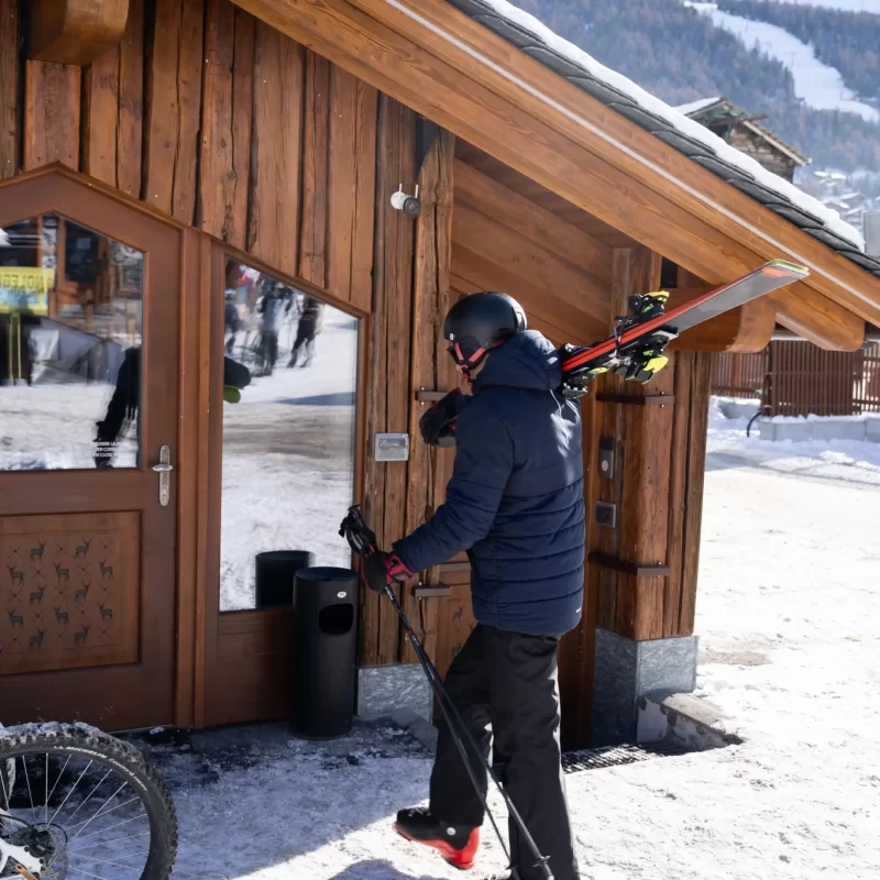 Una persona entra in una baita di legno con sci e bastoncini su una spalla nella neve.